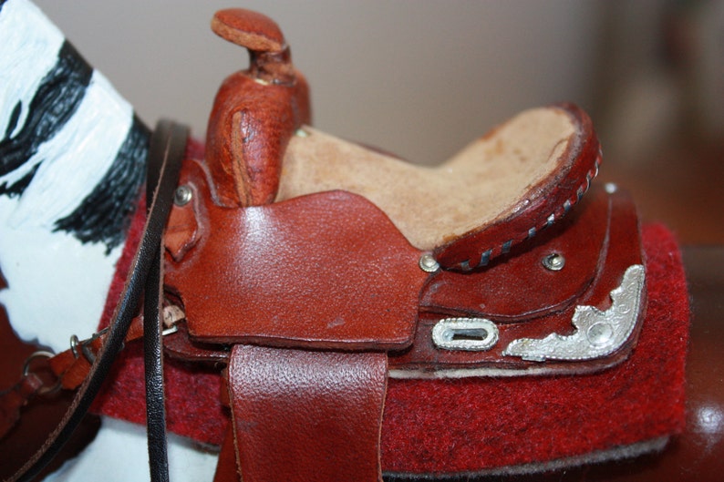 Puede incluir: Una silla de montar de cuero marr&oacute;n con detalles plateados y un asiento de fieltro rojo. La silla est&aacute; unida a un caballo blanco y negro.