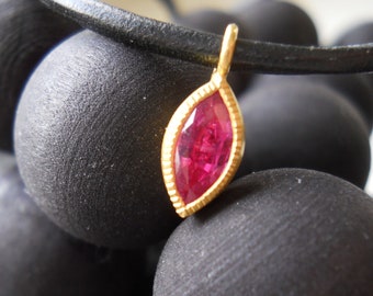 May include: A gold pendant with a red gemstone in the shape of a marquise. The pendant is hanging from a black leather cord and is resting on a pile of black spheres.