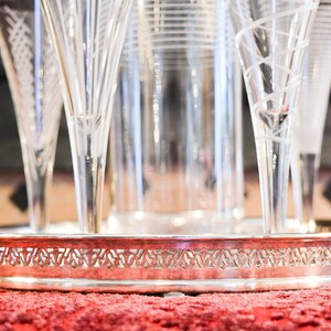 May include: A silver serving tray with a decorative border, holding a set of six clear glass champagne flutes.