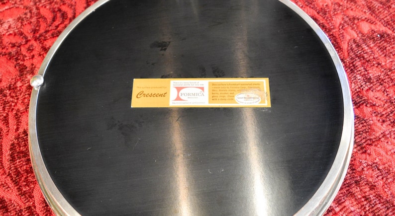 May include: A round, silver-rimmed serving tray with a black Formica surface. The tray has a label that reads "Crescent" and "Formica" with a small logo that says "The Laminates & Decorative Surfacing Division of American Cyanamid Company".