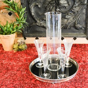 May include: A silver tray with a black surface holds a clear glass pitcher and four clear glass champagne flutes. The tray is on a red textured surface.