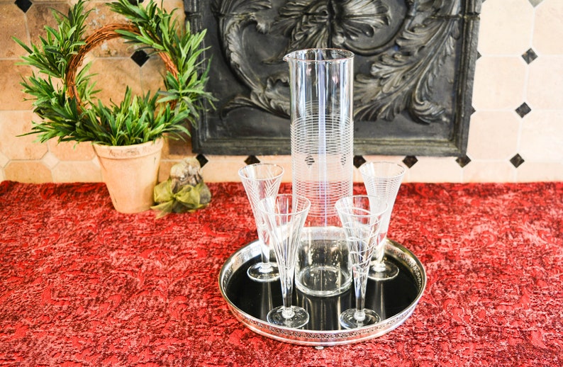 May include: A silver tray with a black surface holds a clear glass pitcher and four clear glass champagne flutes. The tray is on a red textured surface.