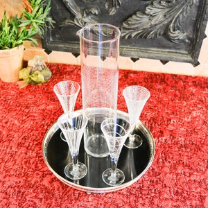 May include: A set of four clear glass champagne flutes and a tall clear glass pitcher with a textured design, all on a black tray with a silver rim. The tray is on a red textured surface.