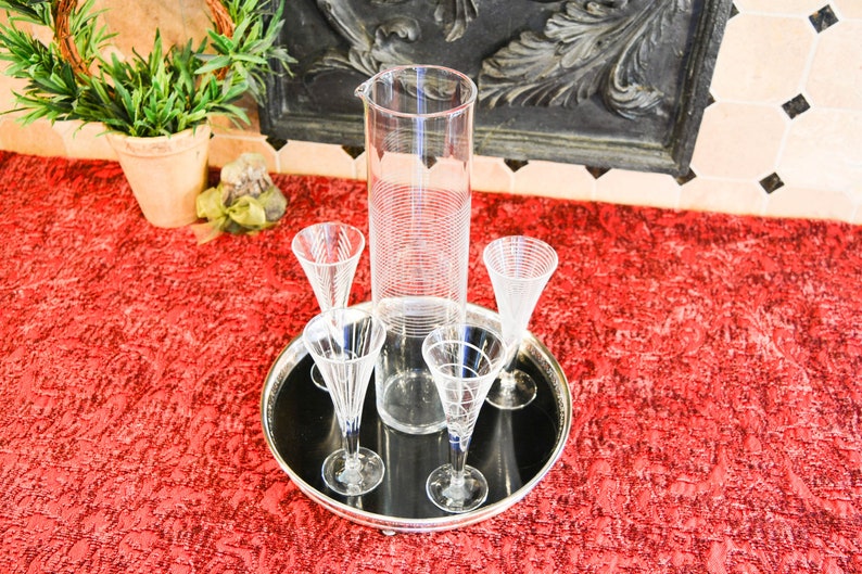 May include: A set of four clear glass champagne flutes and a tall clear glass pitcher with a textured design, all on a black tray with a silver rim. The tray is on a red textured surface.