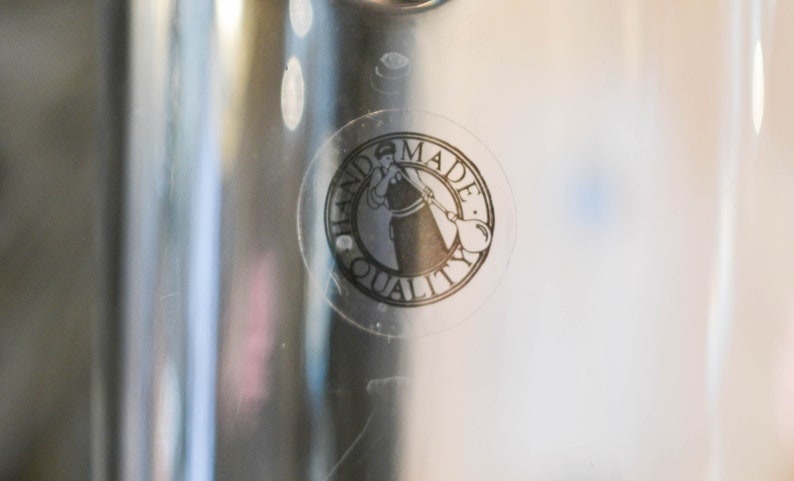 May include: A clear glass bottle with a black and white sticker that says "Handmade Quality" and features a woman blowing glass.