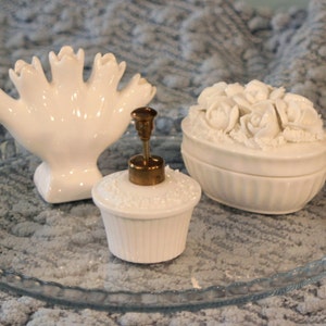 May include: A white porcelain tray with three decorative items: a hand-shaped vase, a floral-shaped trinket box, and a white bottle with a gold top.