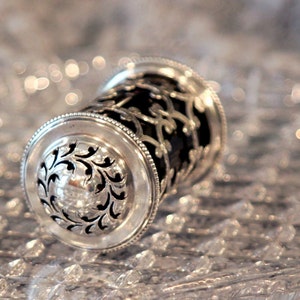 May include: A silver and black decorative object with intricate floral designs. The object is made of metal and has a cylindrical shape.