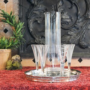 May include: A silver tray with four clear champagne flutes and a tall, clear glass pitcher with a decorative etched pattern. The tray is on a red tablecloth.