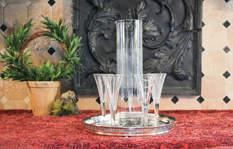 May include: A silver tray with four clear champagne flutes and a tall, clear glass pitcher with a decorative etched pattern. The tray is on a red tablecloth.