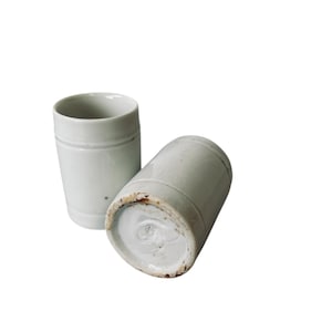 May include: Two off-white ceramic tumblers. One is upright, the other is lying on its side. Each has a cylindrical shape with a slightly flared rim and a subtle horizontal band near the top. The bottom of the tumbler on its side shows a circular design.