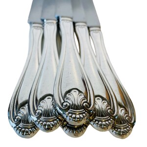 May include: A set of five silver dinner knives with ornate handles. The knives are arranged in a stack with the handles facing up.