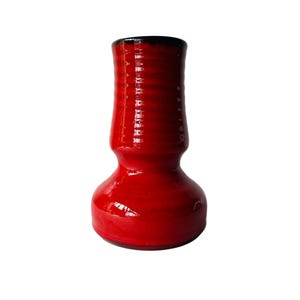 May include: A glossy red ceramic vase with a black rim. The vase has a bulbous base and a cylindrical upper section with horizontal ridges. The vase is a vibrant red colour and appears to be handmade.