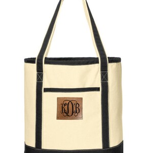 May include: A cream canvas tote bag with black trim and handles. The bag has a brown leather patch with the monogram "KOB" in black.