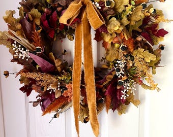 May include: A fall-themed wreath with a variety of leaves in shades of brown, red, and gold. The wreath is adorned with berries and a mustard-colored velvet ribbon. The wreath is displayed on a white door.