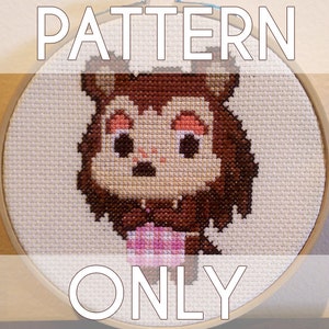 Puede incluir: Patrón de punto de cruz de un personaje de dibujos animados marrón y rosa con ojos grandes y una expresión de sorpresa. El personaje sostiene un objeto rosa. El patrón está enmarcado en un aro de madera.