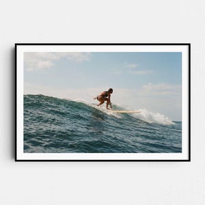 May include: A framed photograph captures a surfer riding a wave. The surfer, in a dark swimsuit, is balanced on a longboard. The wave is a deep blue, with white foam. The sky is a light blue with scattered clouds. The photograph is in a black frame.
