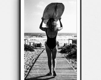May include: Black and white photograph of a person walking on a wooden boardwalk towards the ocean, carrying a surfboard overhead. The person is wearing a black swimsuit. The image is framed in a black border.
