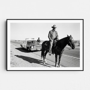 May include: Black and white photograph of a man on horseback next to a police car on a desert highway. A police officer stands near the car. The man on the horse wears a cowboy hat and jacket. The image is framed in a black border.