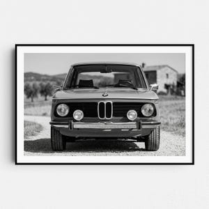 May include: Black and white photograph of a vintage BMW car, viewed head-on. The car has a classic design with round headlights, a prominent front grille, and a chrome bumper. The car is parked on a gravel road with a rural background.