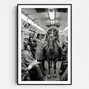 Puede incluir: Fotografía en blanco y negro de un vaquero montado a caballo dentro de un vagón de metro. Los pasajeros están sentados y de pie, algunos mirando al vaquero. El vagón de metro tiene grafitis en las paredes.