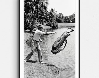 May include: Black and white framed print of a golfer throwing a golf bag into a lake. The golfer is wearing a visor, collared shirt, and slacks. Palm trees and a golf course are in the background.