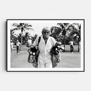 May include: Black and white photograph of a person carrying two golf bags filled with golf clubs. The person is wearing sunglasses and a white jumpsuit, walking on a golf course. Palm trees and other golfers are visible in the background.