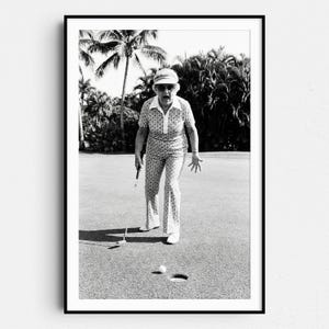 Könnte beinhalten: Schwarz-Weiß-Druck einer Frau beim Golfspielen. Sie trägt ein gemustertes Outfit, einen Visor und eine Sonnenbrille, mit offenem Mund vor Überraschung. Ein Golfball liegt in der Nähe des Lochs, und sie hält einen Golfschläger.