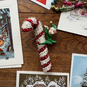May include: A decorative candy cane ornament featuring alternating diagonal stripes of red sequins and white iridescent beads. A small, green-clad elf figure with a pointed hat and painted facial features is attached to the side of the candy cane.