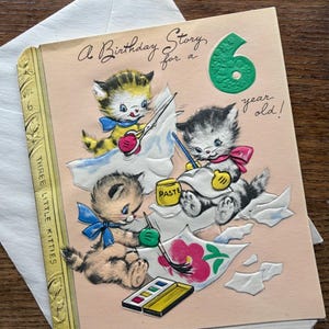 Puede incluir: Tarjeta de cumplea&ntilde;os vintage con una portada rosa que muestra tres gatitos pintando. La tarjeta dice "A Birthday Story for a 6 year old!". Los gatitos est&aacute;n representados con pinceles, pintura y pegamento. La tarjeta tiene forma de libro.