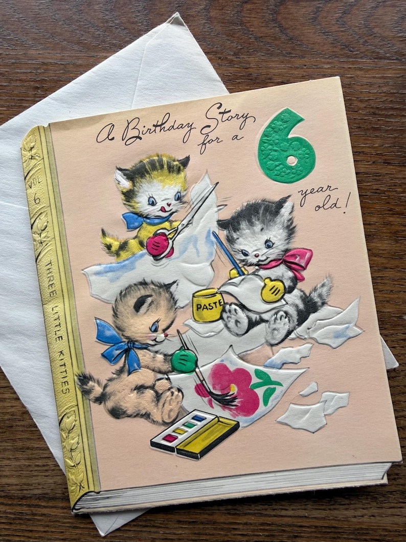 Puede incluir: Tarjeta de cumplea&ntilde;os vintage con una portada rosa que muestra tres gatitos pintando. La tarjeta dice "A Birthday Story for a 6 year old!". Los gatitos est&aacute;n representados con pinceles, pintura y pegamento. La tarjeta tiene forma de libro.