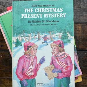 May include: A book cover with a green background and the title "The Christmas Present Mystery" by Marion M. Markham. The cover features an illustration of two girls in pink jackets and red and white striped scarves standing in a snowy forest.
