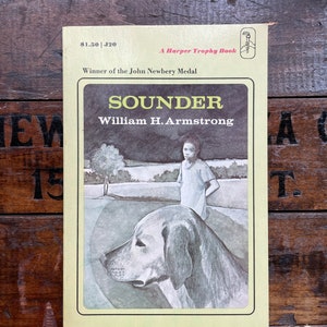 May include: A yellow paperback book cover with the title "Sounder" by William H. Armstrong. The book is a Harper Trophy Book and a winner of the John Newbery Medal. The cover features a black and white illustration of a young girl standing next to a large dog.