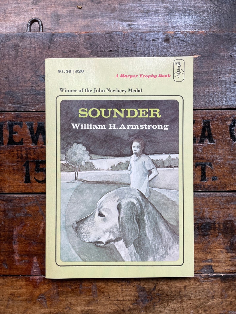 May include: A yellow paperback book cover with the title "Sounder" by William H. Armstrong. The book is a Harper Trophy Book and a winner of the John Newbery Medal. The cover features a black and white illustration of a young girl standing next to a large dog.