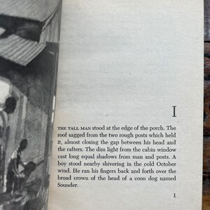 May include: A black and white photo of a book with the text "THE TALL MAN stood at the edge of the porch. The roof sagged from the two rough posts which held it, almost closing the gap between his head and the rafters. The dim light from the cabin window cast long equal shadows from man and posts. A boy stood nearby shivering in the cold October wind. He ran his fingers back and forth over the broad crown of the head of a coon dog named Sounder."