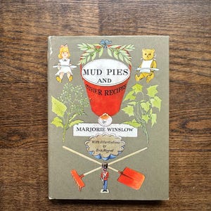 May include: A vintage book titled "Mud Pies and Other Recipes" by Marjorie Winslow. The cover features illustrations of a girl, a teddy bear, gardening tools, and foliage. The book has a green background with red and white text.