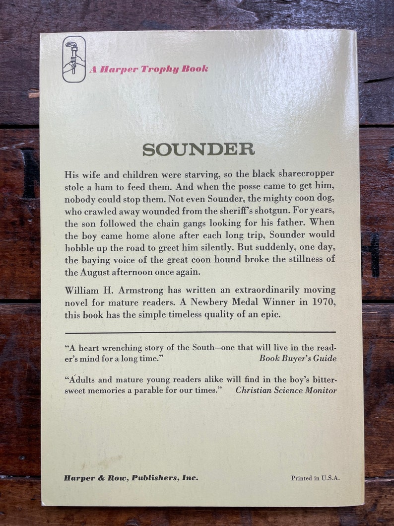 May include: A vintage book cover with a yellow background and brown text. The title of the book is "Sounder" and the author is William H. Armstrong. The book is a Newbery Medal Winner from 1970. The back cover includes quotes from Book Buyer's Guide and Christian Science Monitor.