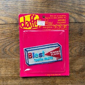 May include: A rectangular embroidered patch featuring a white toothpaste tube design with blue borders. The text reads Blest TOOTH PASTE and HEAVENLY FLAVOR in red and blue. It is mounted on a bright pink cardboard backing with the brand name Daffy.