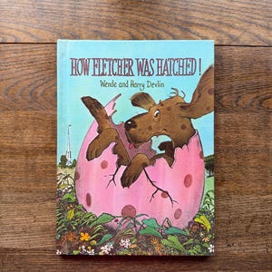 May include: A children's book titled "How Fletcher Was Hatched!" features a brown dog emerging from a pink egg. The cover has a light blue background with the title in red and the authors' names. The illustration includes green foliage and a church.