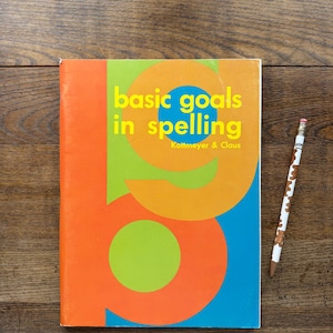 May include: A colorful book titled "basic goals in spelling" with a large orange and green design. A pencil with a bear design rests on a wooden surface next to the book. The book's cover is orange, green, and blue.
