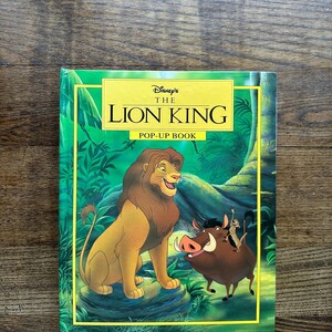 May include: A hardcover pop-up book titled "Disney's The Lion King." The cover features illustrations of Simba, Pumbaa, Timon, and a jungle background. The book has a green border and a yellow title.