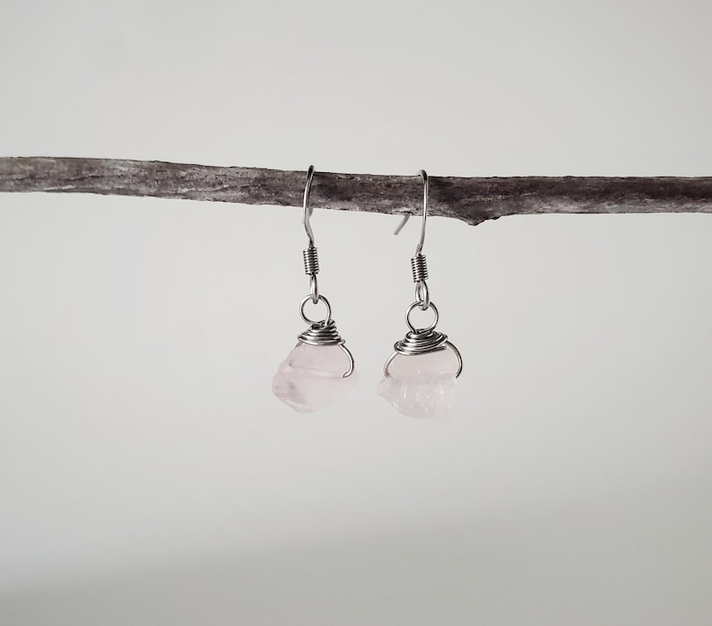May include: A pair of rose quartz earrings. Each earring features a raw, light pink rose quartz stone wrapped in silver-colored wire, suspended from a silver-colored ear hook. The earrings are displayed against a neutral background.