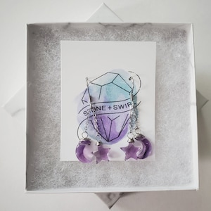May include: A pair of amethyst earrings featuring a crescent moon and star design. The earrings are suspended from silver-colored hooks and chains. The earrings are displayed in a white gift box with a card that reads "STONE + SWIRL".