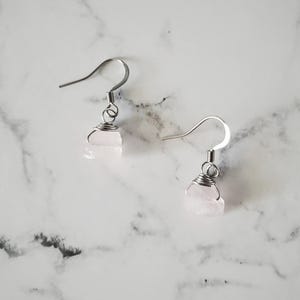 May include: A pair of earrings featuring raw, light pink rose quartz stones. The stones are wrapped in silver-colored wire and suspended from silver-colored ear wires. The earrings are displayed on a white and gray marble surface.