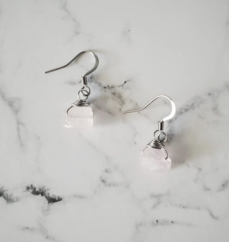 May include: A pair of earrings featuring raw, light pink rose quartz stones. The stones are wrapped in silver-colored wire and suspended from silver-colored ear wires. The earrings are displayed on a white and gray marble surface.