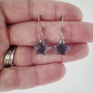 Op de afbeelding: Een paar hangende oorbellen met gefacetteerde, stervormige blauwgrijze stenen bedels die aan zilverkleurige metalen vishaken hangen. Elke sterbedel is aan de haak bevestigd met een kleine zilverkleurige ring en gewikkeld metaaldraad.