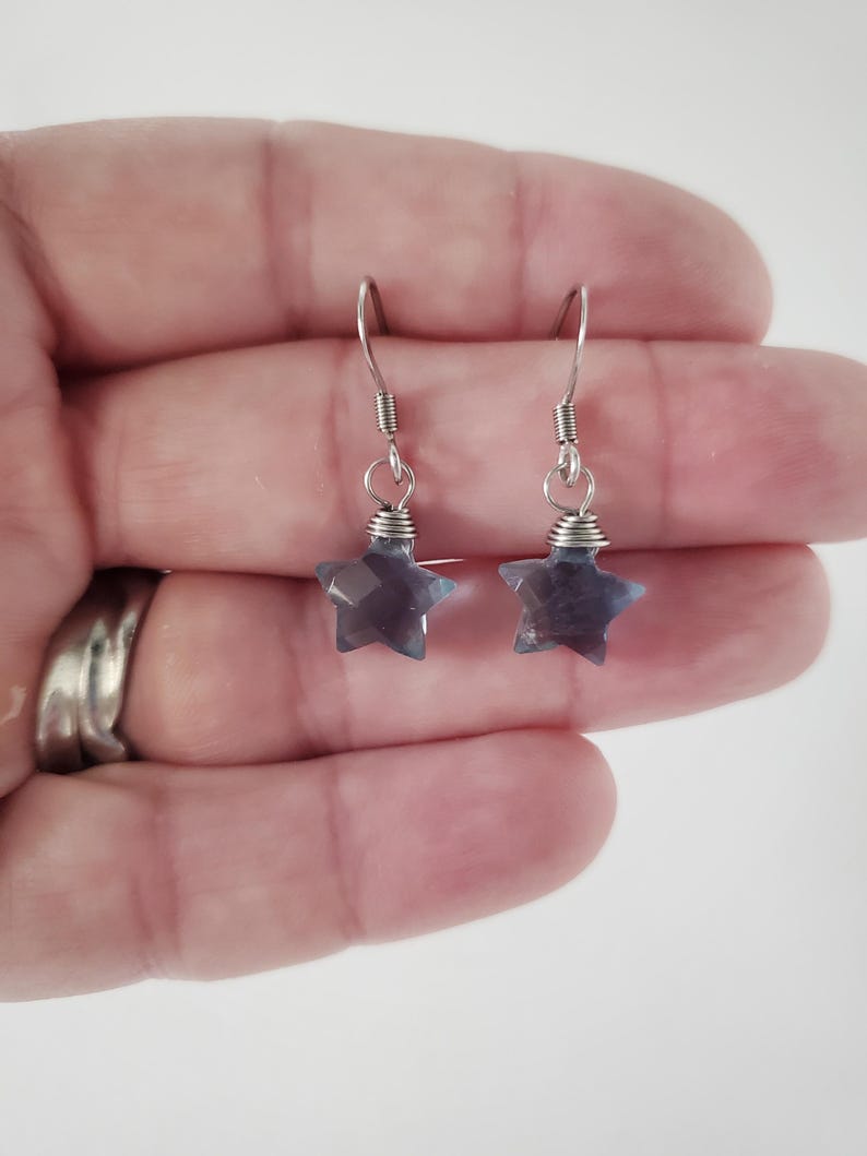 Op de afbeelding: Een paar hangende oorbellen met gefacetteerde, stervormige blauwgrijze stenen bedels die aan zilverkleurige metalen vishaken hangen. Elke sterbedel is aan de haak bevestigd met een kleine zilverkleurige ring en gewikkeld metaaldraad.