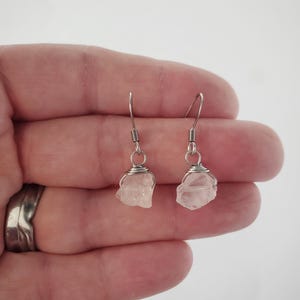 May include: A pair of earrings featuring raw, light pink rose quartz stones wrapped in silver-colored wire. The earrings have a simple hook design for pierced ears, showcasing the natural beauty of the crystals. The earrings are held in a hand.