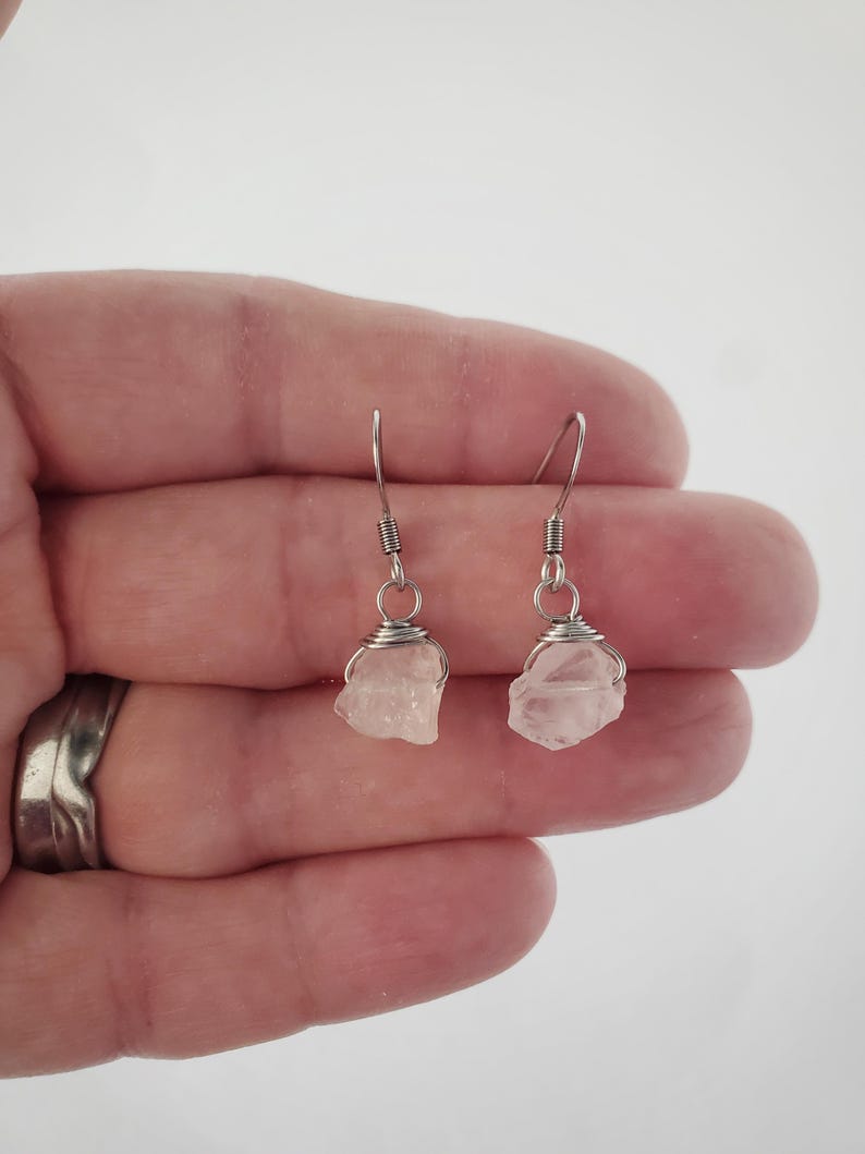 May include: A pair of earrings featuring raw, light pink rose quartz stones wrapped in silver-colored wire. The earrings have a simple hook design for pierced ears, showcasing the natural beauty of the crystals. The earrings are held in a hand.