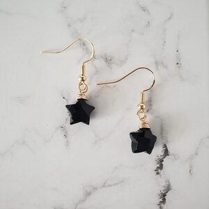 May include: A pair of gold-colored dangle earrings. Each earring features a faceted black star pendant, suspended from a gold-colored hook. The star is wire-wrapped with gold-colored wire. The background is a white and gray marble pattern.