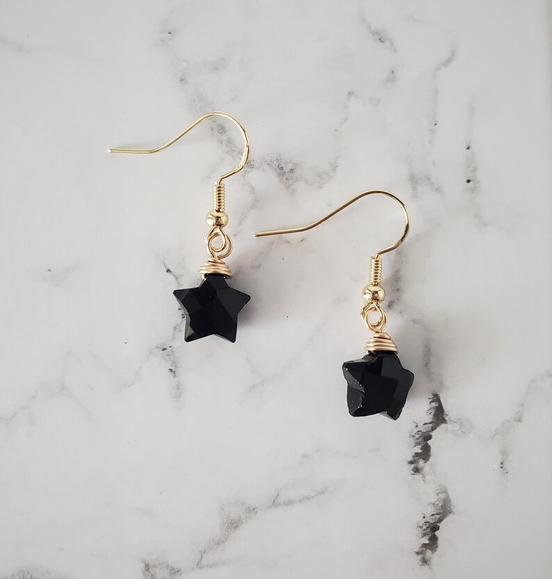 May include: A pair of gold-colored dangle earrings. Each earring features a faceted black star pendant, suspended from a gold-colored hook. The star is wire-wrapped with gold-colored wire. The background is a white and gray marble pattern.